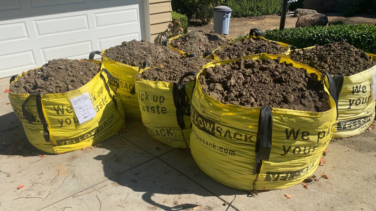 A Medium Yellowsack filled with soil, a planter bed behind it